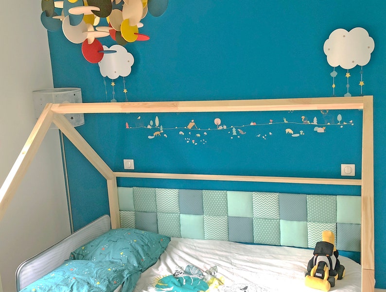 Puede incluir: Una cama con estructura de madera y cabecero acolchado. La pared es de color azul turquesa. El cabecero est&aacute; hecho de un mosaico de cuadrados de tela azul claro y gris. Un m&oacute;vil de nube blanca cuelga del techo.
