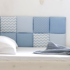 May include: A white bed with a blue and white patterned headboard. The headboard is made up of individual squares with different patterns, including stars, stripes, and zigzags. A grey blanket is folded on the bed.