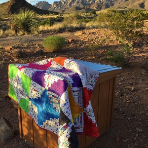 May include: A colorful handmade quilt with a starburst design, draped over a wooden structure. The quilt features vibrant blocks of green, blue, purple, orange, and red, set against a white background. The scene is set outdoors with a mountain backdrop.