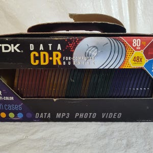 May include: A box of TDK Data CD-Rs in multi-color slim cases. The box is black with red and yellow accents and features the text "CD-R" and "50 Pack". The CDs are visible through a window in the box.