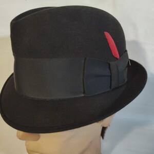 May include: A black fedora hat with a wide black band and a black bow. A red feather is tucked into the side of the hat. The hat is displayed on a mannequin head.