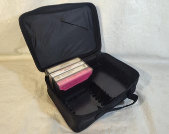 May include: A black zippered carrying case for cassette tapes with a handle. The interior features molded plastic slots for organizing tapes, with three cassette tapes currently stored inside, including titles like Sweet Caroline and Greatest Hits.