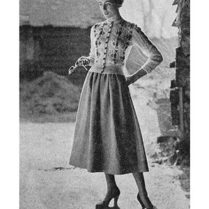 May include: A black and white photo of a woman wearing a white knitted sweater with hand embroidery and a matching skirt. The sweater has a button-up front and a round neckline. The skirt is a-line and falls to just below the knee. The woman is standing in front of a brick wall.