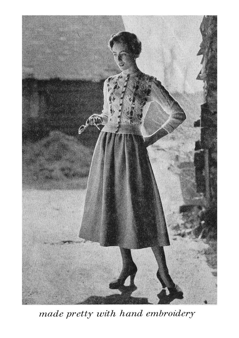 May include: A black and white photo of a woman wearing a white knitted sweater with hand embroidery and a matching skirt. The sweater has a button-up front and a round neckline. The skirt is a-line and falls to just below the knee. The woman is standing in front of a brick wall.