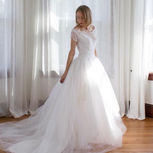 May include: A white wedding dress with a tulle skirt and a lace bodice. The dress is being worn by a person in a room with white curtains.