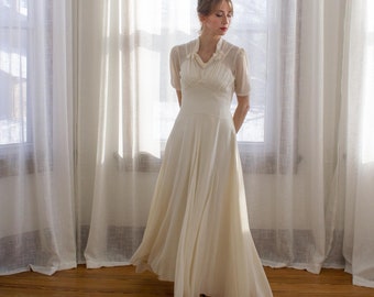 May include: A woman in a white vintage wedding dress stands in front of a window with sheer curtains. The dress has a fitted bodice with a ruffled neckline and a flowing skirt.