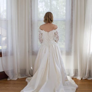 May include: A white wedding dress with a long train. The dress features a lace bodice with long sleeves and a full skirt. The bride is standing in front of a window with sheer white curtains.