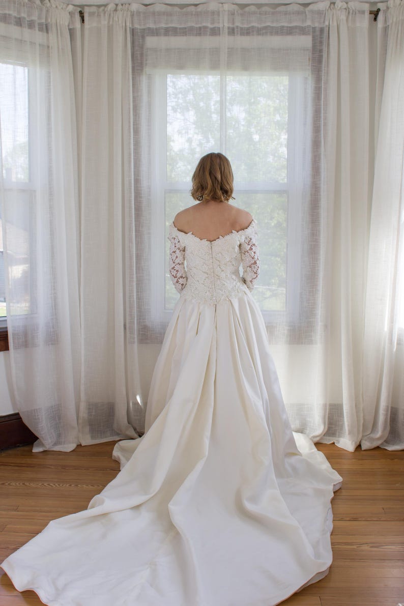 May include: A white wedding dress with a long train. The dress features a lace bodice with long sleeves and a full skirt. The bride is standing in front of a window with sheer white curtains.