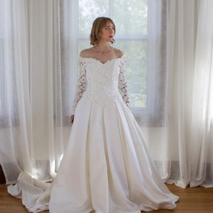 May include: A white off-the-shoulder wedding dress with long lace sleeves and a full skirt. The bodice features intricate lace detailing. The dress is displayed in front of a window with sheer white curtains and a wooden floor.