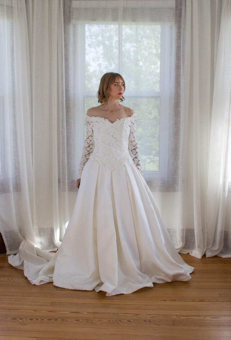 May include: A white off-the-shoulder wedding dress with long lace sleeves and a full skirt. The bodice features intricate lace detailing. The dress is displayed in front of a window with sheer white curtains and a wooden floor.