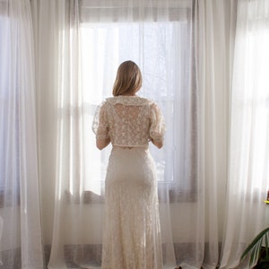 May include: A woman in a white lace dress stands in front of a window with white curtains. The dress has a fitted bodice and a flowing skirt. The woman is looking out the window.