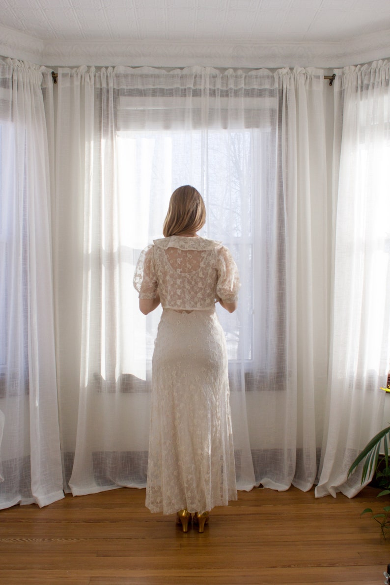 May include: A woman in a white lace dress stands in front of a window with white curtains. The dress has a fitted bodice and a flowing skirt. The woman is looking out the window.