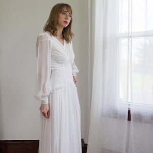 May include: A woman wearing a white, long-sleeved, lace dress with a ruffled skirt. She is standing in a room with a window and white curtains.
