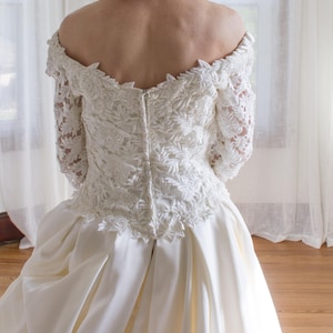 May include: A white wedding dress with an off-the-shoulder neckline and long lace sleeves. The bodice features intricate floral embroidery, and the skirt is made of smooth, flowing fabric. The dress is designed for a formal occasion.