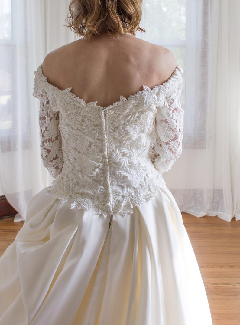 May include: A white wedding dress with an off-the-shoulder neckline and long lace sleeves. The bodice features intricate floral embroidery, and the skirt is made of smooth, flowing fabric. The dress is designed for a formal occasion.