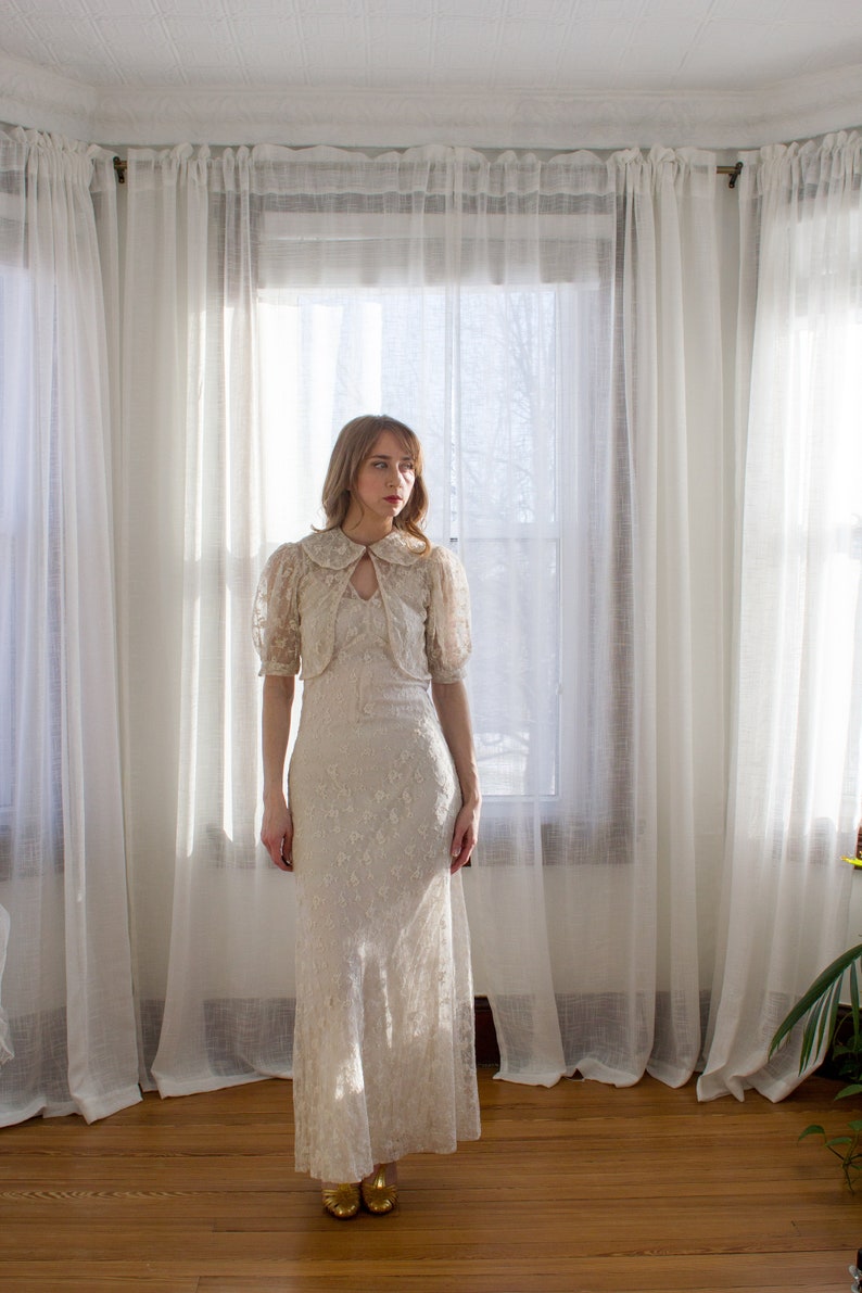 May include: A woman wearing a white lace dress with a high collar and puff sleeves. She is standing in front of a window with white curtains.