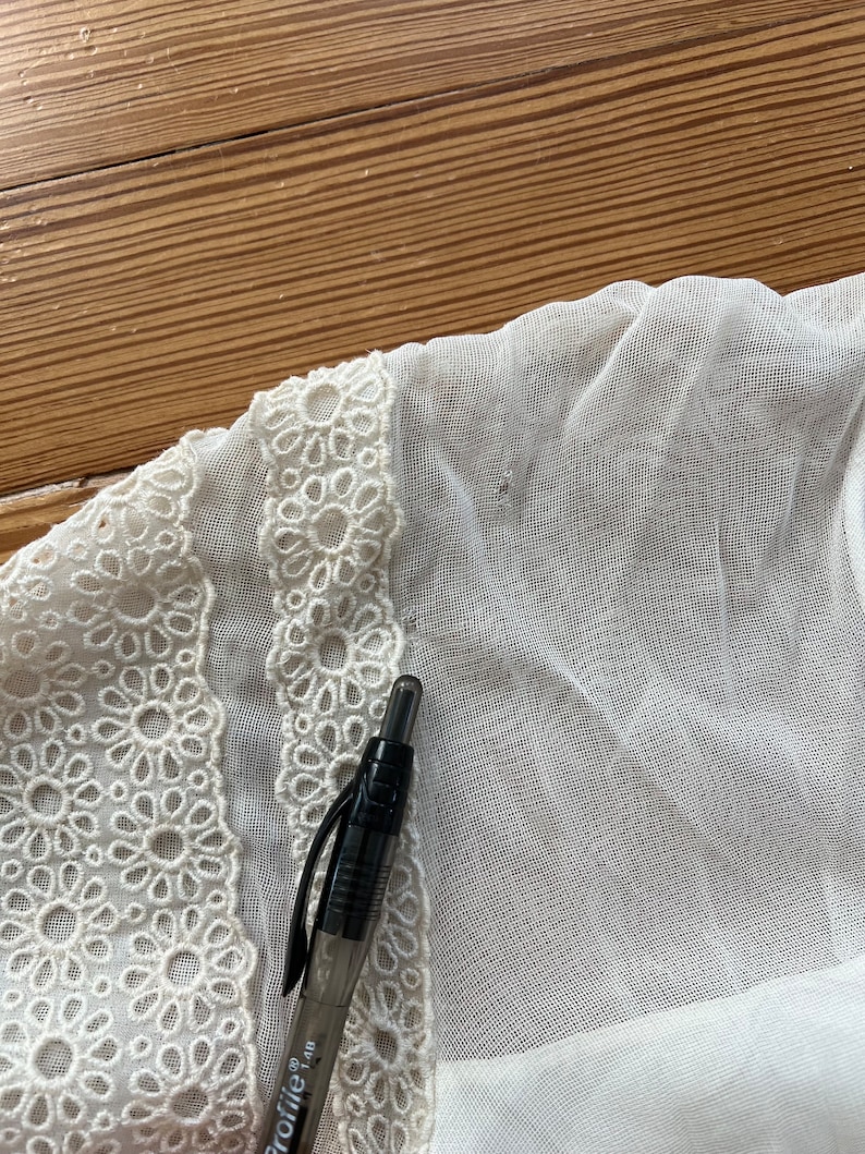 Pode incluir: Tecido branco transparente com uma borda de renda branca. Uma caneta preta est&aacute; sobre o tecido.