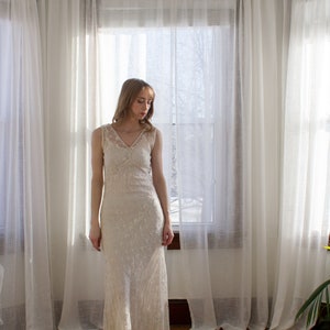 May include: A woman in a white lace dress stands in front of a window with sheer curtains. The dress has a V-neckline and a fitted silhouette.