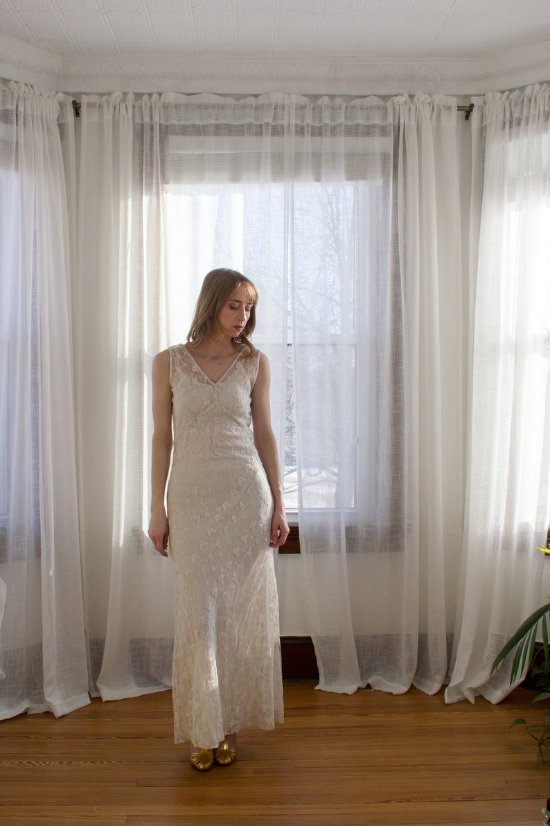 May include: A woman in a white lace dress stands in front of a window with sheer curtains. The dress has a V-neckline and a fitted silhouette.