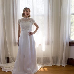 May include: A white wedding dress with a lace bodice and a flowing skirt. The dress is worn by a woman standing in front of a window with white curtains.