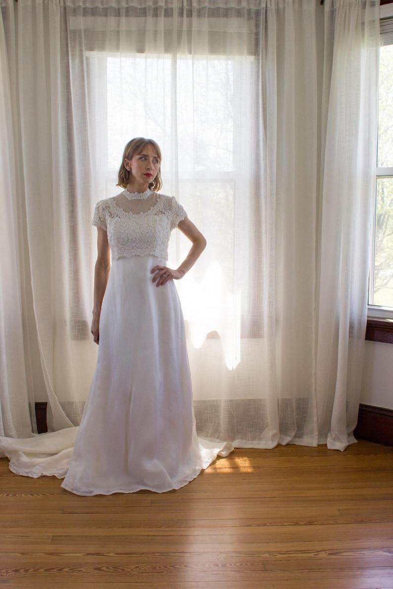 May include: A white wedding dress with a lace bodice and a flowing skirt. The dress is worn by a woman standing in front of a window with white curtains.