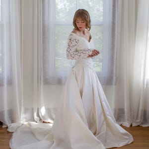 May include: A vintage ivory wedding dress with a long train. The dress features a lace bodice with long sleeves and a V-neckline. The skirt is full and flowing, made of a smooth, satin-like fabric. The bride is standing in front of a window.
