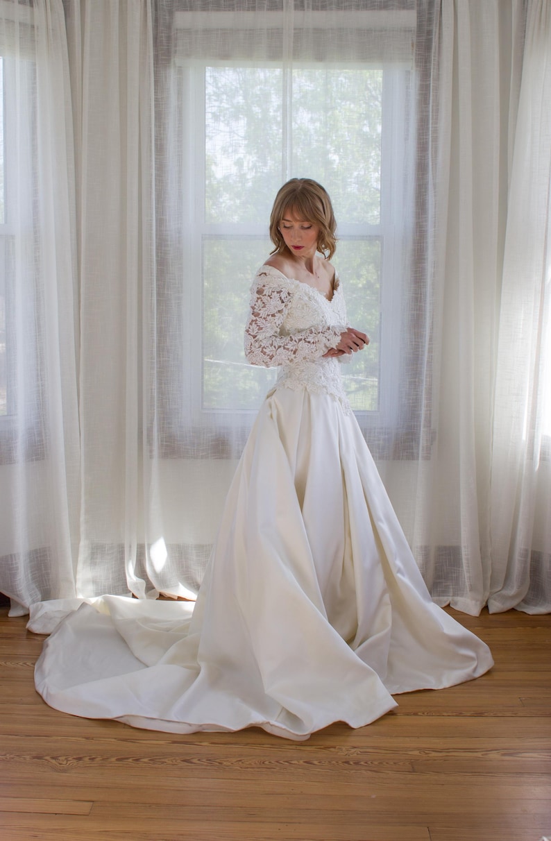May include: A vintage ivory wedding dress with a long train. The dress features a lace bodice with long sleeves and a V-neckline. The skirt is full and flowing, made of a smooth, satin-like fabric. The bride is standing in front of a window.