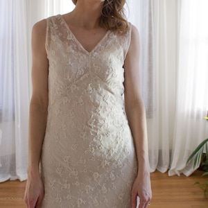 May include: A white sleeveless lace dress with a V-neckline. The dress is made of delicate floral lace and has a fitted silhouette.