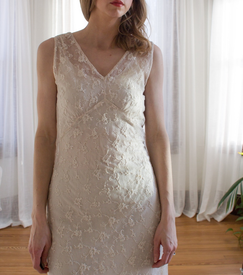 May include: A white sleeveless lace dress with a V-neckline. The dress is made of delicate floral lace and has a fitted silhouette.