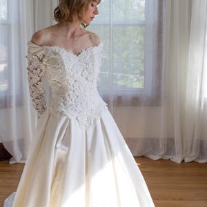 May include: A white off-the-shoulder wedding dress with a lace bodice and long sleeves. The skirt is a full, flowing satin. The dress is in front of a window with sheer white curtains and a wooden floor.