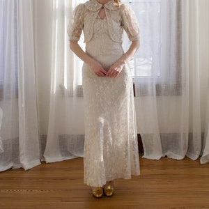 May include: A white lace dress with a matching lace jacket. The dress has a fitted bodice and a flowing skirt. The jacket has a Peter Pan collar and short sleeves.