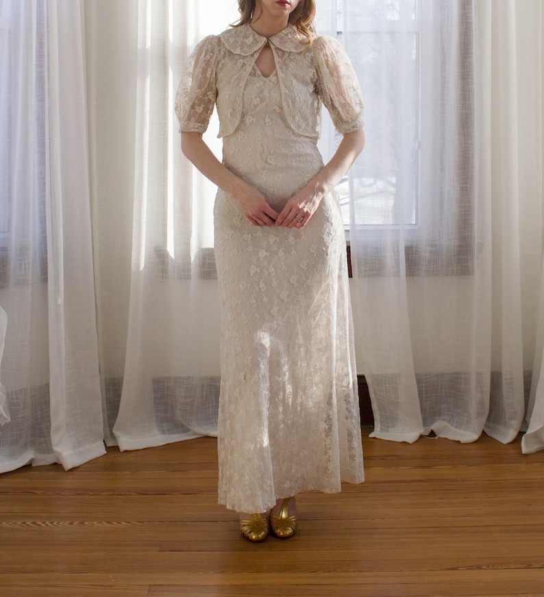 May include: A white lace dress with a matching lace jacket. The dress has a fitted bodice and a flowing skirt. The jacket has a Peter Pan collar and short sleeves.