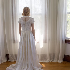 May include: A woman in a white lace wedding dress stands in front of a window with sheer curtains. The dress has a fitted bodice and a flowing skirt. The woman is looking out the window.