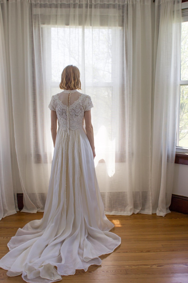 May include: A woman in a white lace wedding dress stands in front of a window with sheer curtains. The dress has a fitted bodice and a flowing skirt. The woman is looking out the window.
