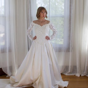 May include: A white wedding dress with a lace bodice and long sleeves. The off-the-shoulder neckline is adorned with delicate lace. The skirt is full and flowing, with a train that extends behind the dress. The dress is made of a satin-like material.