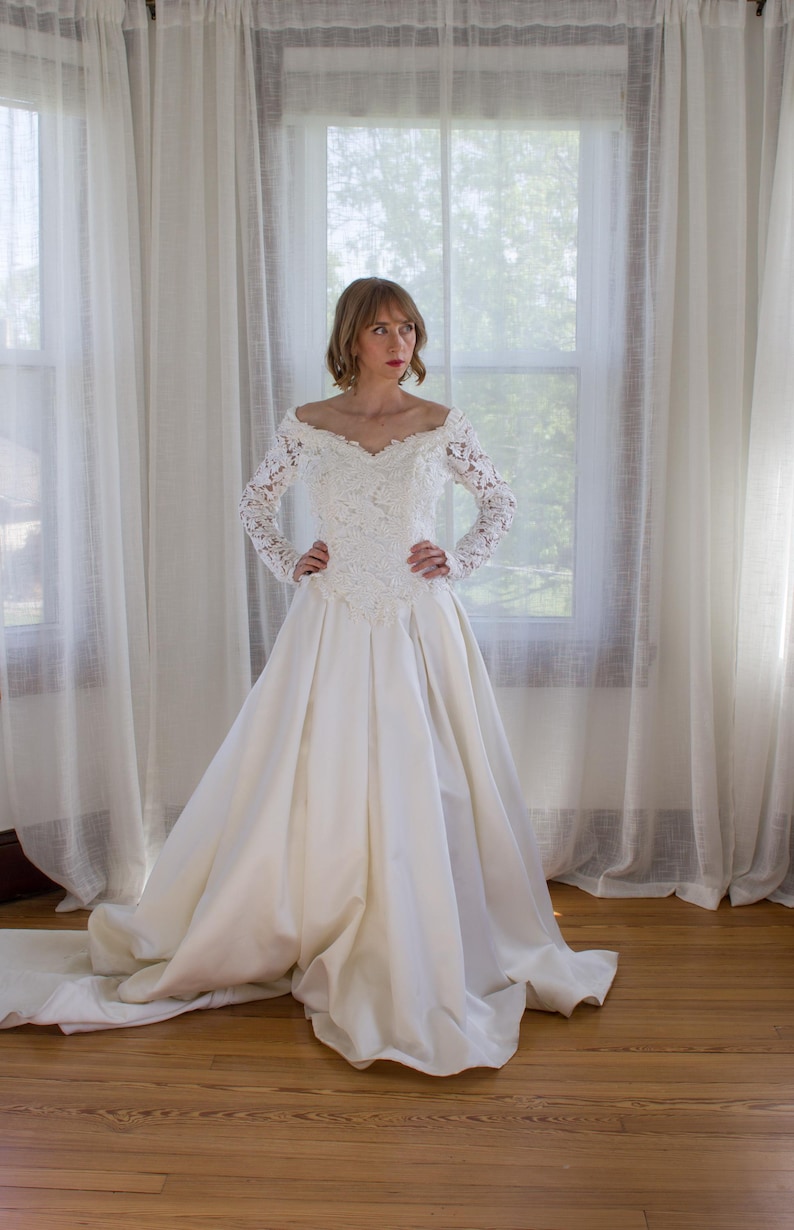 May include: A white wedding dress with a lace bodice and long sleeves. The off-the-shoulder neckline is adorned with delicate lace. The skirt is full and flowing, with a train that extends behind the dress. The dress is made of a satin-like material.