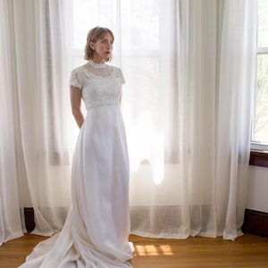 May include: A white wedding dress with a lace bodice and a flowing skirt. The dress has a high neckline and short sleeves. The bride is standing in front of a window with white curtains.