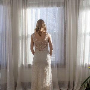 May include: A woman in a white lace dress stands in front of a window with white curtains. She is looking out the window.
