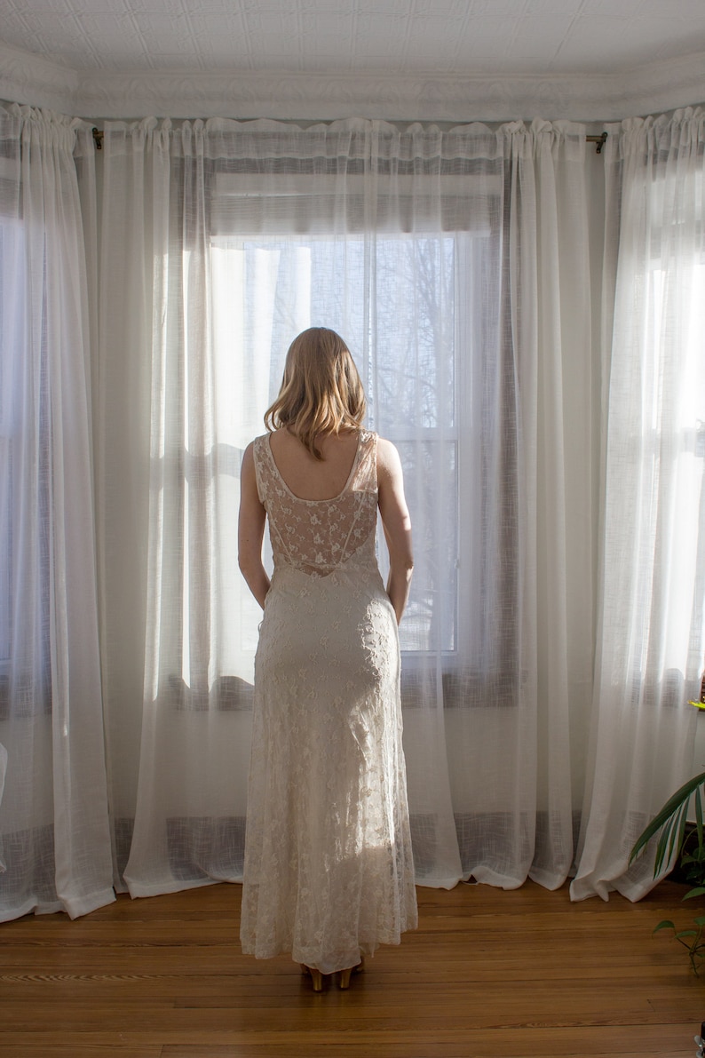 May include: A woman in a white lace dress stands in front of a window with white curtains. She is looking out the window.
