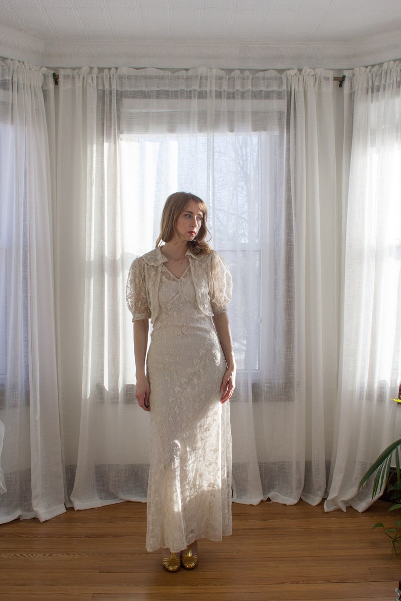 May include: A woman wearing a white lace dress with a matching lace jacket. She is standing in front of a window with white curtains.