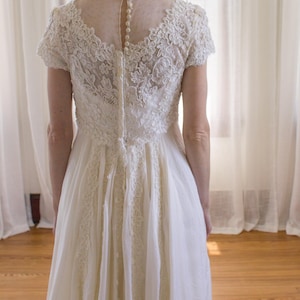 May include: A white lace wedding dress with a sheer back and a row of buttons down the center. The dress has a fitted bodice and a flowing skirt.