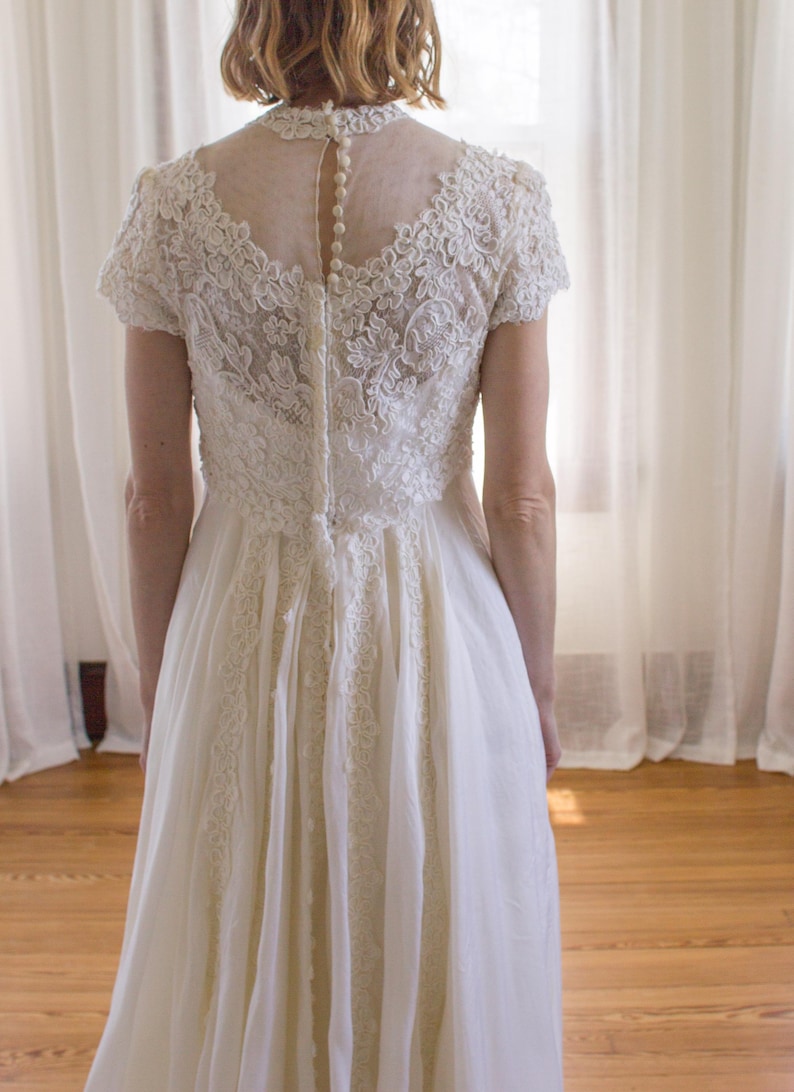 May include: A white lace wedding dress with a sheer back and a row of buttons down the center. The dress has a fitted bodice and a flowing skirt.