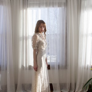 May include: A woman wearing a white lace dress with a long skirt and a fitted bodice. She is standing in a room with white curtains and a window behind her.