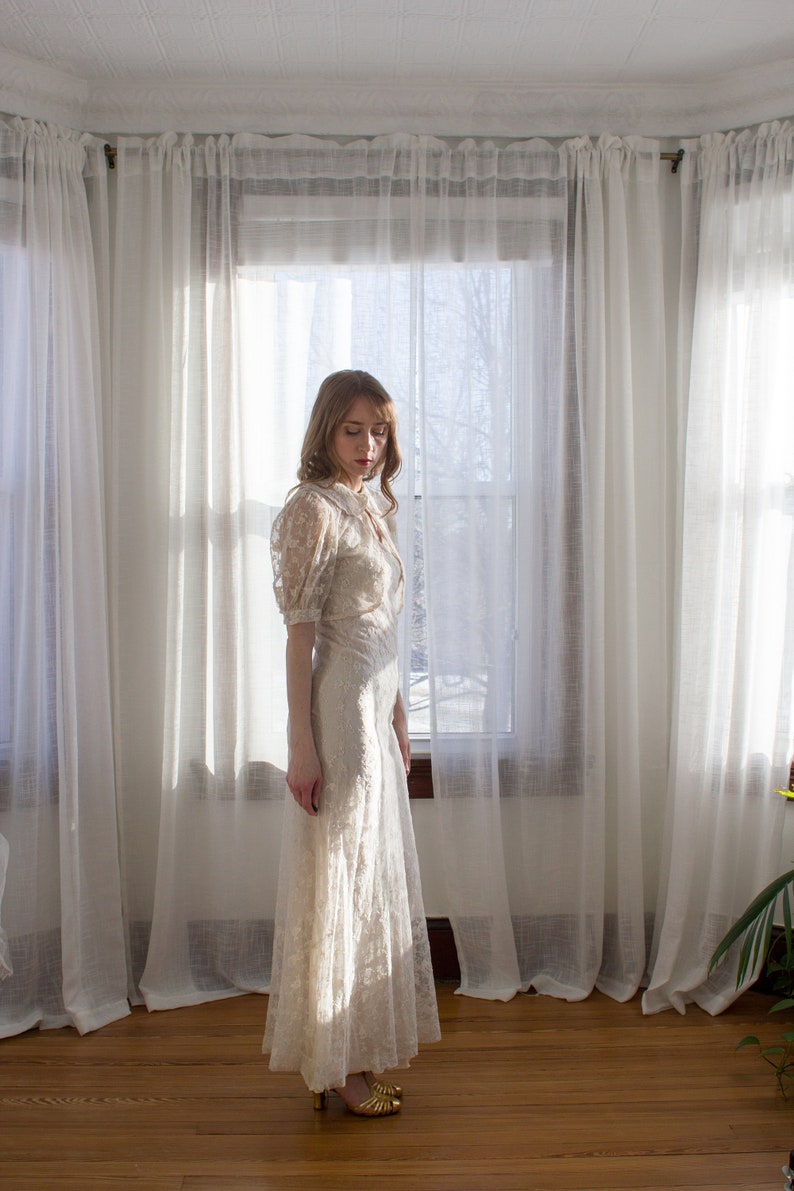 May include: A woman wearing a white lace dress with a long skirt and a fitted bodice. She is standing in a room with white curtains and a window behind her.