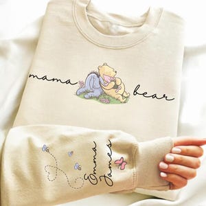 Puede incluir: Sudadera color crema con un gráfico de Winnie the Pooh y Eeyore con las palabras "mama bear". La manga muestra los nombres "Emma James" con abejas y un diseño de mariposa.