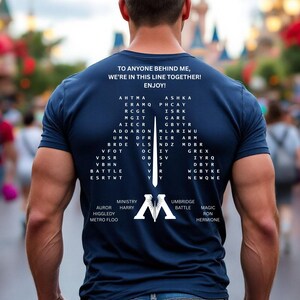 May include: A navy blue short-sleeved t-shirt featuring white text on the back. The text reads: TO ANYONE BEHIND ME, WE'RE IN THIS LINE TOGETHER! ENJOY! Below this, a large letter M is formed by columns of letters, with words like MINISTRY, HARRY, UMBRIDGE, BATTLE, MAGIC, RON, and HERMIONE printed at the bottom.