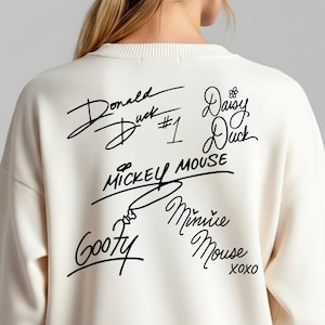 Puede incluir: Sudadera color crema con firmas negras de personajes de Disney: Pato Donald, Daisy, Mickey Mouse, Minnie Mouse y Goofy. También está presente el texto "#1". La sudadera es de estilo casual.