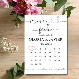 Può includere: Un invito di nozze bianco con il testo "reserva la fecha" e "para la boda de GLORIA & JAVIER JUNIO 2026". Un cuore rosa evidenzia il primo giorno del mese. L'invito è decorato con fiori rosa e verde.