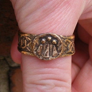 Könnte beinhalten: Ein goldfarbener Ring mit einem keltischen Knotendesign und einem zentralen Motiv aus drei stilisierten Kreisen. Der Ring wird am Daumen getragen.