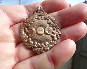 May include: A bronze pendant with a triple moon design and a leafy border. The pendant is inscribed with the word "DOC".